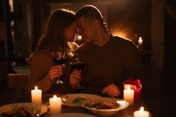 Happy young couple in love hugging holding glasses having romantic dinner date with candles in dark light, drinking wine, celebrating Valentines day at home