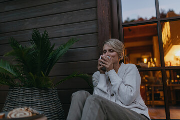 Low angle of woman sitting on the terrace during autumn dusk, sipping warm tea and enjoying the...