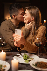 Portrait of happy young couple making selfie on smartphone celebrating Valentine's Day during romantic dinner in restaurant or at home. Happy moments