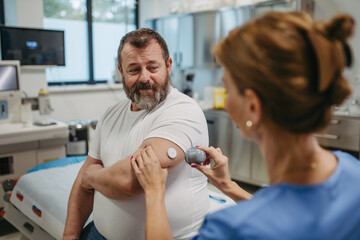 Doctor applying a continuous glucose monitor sensor on patient's arm. Obese, overweight man is at...