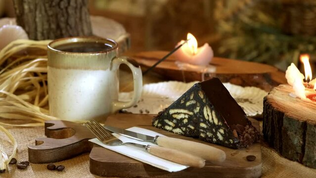 A video in horizontal frame, depicting a delectable mosaic cake on a wicker sackcloth and wooden cutting board. Accompanied by a cup of steaming coffee, burning candles, and incense in the background