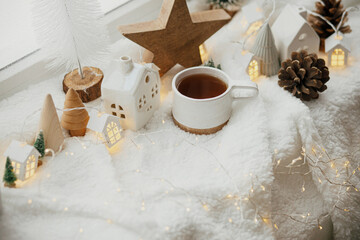 Winter hygge. Stylish cup of tea with modern cute christmas houses, pine cone, wooden star and tree, golden lights on soft warm blanket on windowsill. Christmas cozy still life. Merry Christmas!