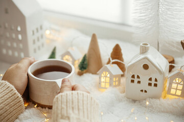 Fototapeta na wymiar Cozy winter. Hands in sweater holding stylish cup of tea with modern christmas decoration, pine cone, wooden star and tree, golden lights on soft warm blanket on windowsill