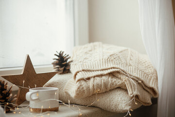 Winter and autumn hygge. Stylish cup of tea with cozy knitted sweater, pine cone, wooden star and golden lights on windowsill in festive scandinavian room. Cozy Christmas. Happy Holidays!