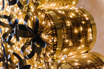 Christmas tree with garlands and gifts in background. Christmas gifts with gold bow. Many boxes with surprises. Happy New Year 2024 and Marry Christmas holiday. Festive glowing golden tree. Closeup.