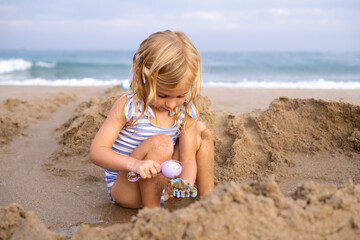 Cute laughing blonde three years old girl playing with sand at beach near sea in Spain.Child activity, happiness.Kid having fun at resort on vacation.
