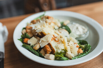 A white bowl full of Cezar salad with croutons, chicken and parmesan cheese. Cezar salad on white plate over wooden table