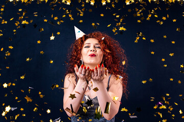 beautiful young red -haired curly woman celebrating birthday or new year and chrismas party while blowing confetti decorations to camera isolated over black background