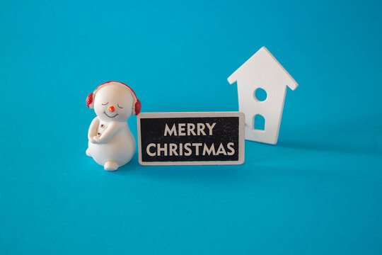Merry Christmas Concept. Merry Christmas text composed on wooden board on beautiful blue background. happy jolly snowman near miniature houses.