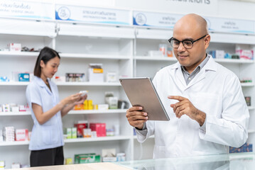 Naklejka na ściany i meble Happy handsome asian male pharmacist wearing eyeglasses and lab coat standing with tablet with coworker. He feels good, trustworthy and proud of his work in the pharmacy drugstore.