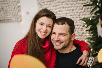 Couple embraces near Christmas tree decorated garlands in interior room. Man hugs woman enjoying winter time together on holiday at home. Happy New Year and Merry Christmas. Closeup love with bokeh.