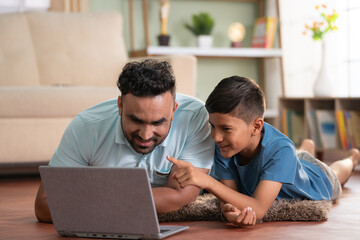 Joyful indian father with son watching laptop by lying on floor at home - concept of cyberspace,...