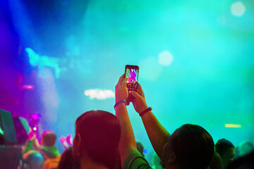 Hands of young man holding smartphone, taking photos, music festival concert, bright neon light. Blurred background