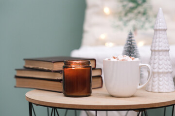 Christmas home decorations - burning candle, books, cup of cocao with marshmellow, ceramic christmas tree on coffee table on green background.