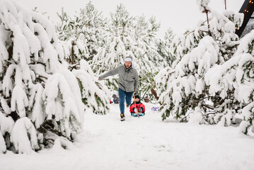 Family walks during snowfall in park. Dad pulling little son on winter day. Children are rolling down hill on sledge in forest. Happy funny child with father ride sled on snowy road in mountains.