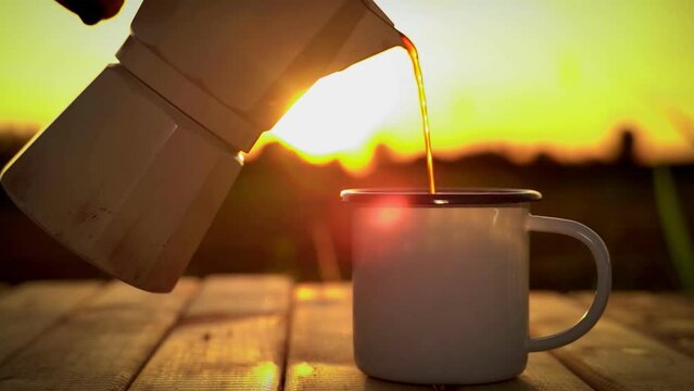 Pour hot coffee from the moka pot. Place a white enamel coffee mug on a wooden table against the backdrop of the morning sunrise.