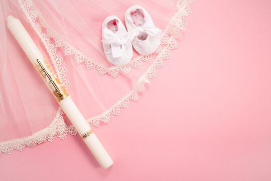 Christening background with baptism baby shoes, dress and candle on pink background.