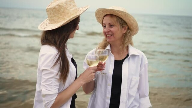 Two young and stylish hipster women wearing trendy summer shirts and hats enjoying wine during their seaside picnic. Attractive girlfriends females feel happy at holiday trip in tropical sea island