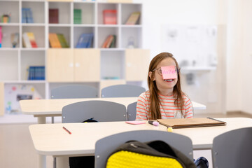 The girl sitting at the class awith a sticky note on her face