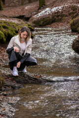 A woman in white jacket and black pants is crouching on a rock in a forest stream