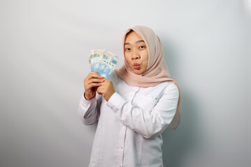 Enthusiastic Asian hijab woman looking excited at camera and holding money rupiah in hand isolated on white background