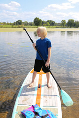 A happy little boy child is learning to stand up paddle board on the lake on a summer day.