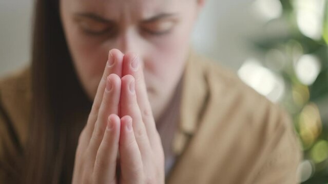 Close up of young caucasian woman praying to God during hard times. Depressed girl seeking divine help in prayer. Female sitting on couch suffering alone with hands together. Selective focus