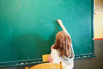 Little schoolgirl writes on the school blackboard with chalk in the classroom - obrazy, fototapety, plakaty