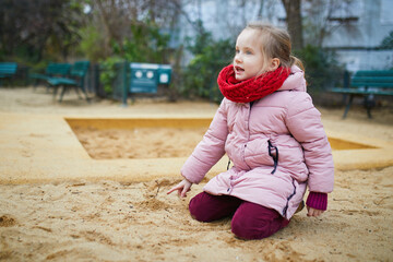 Adorable preschooler girl having fun on playground in sandpit on winter day