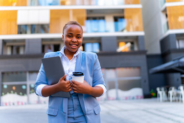 A young business woman with short cropped hair carries a laptop and coffee in her hand in front of a business building - obrazy, fototapety, plakaty