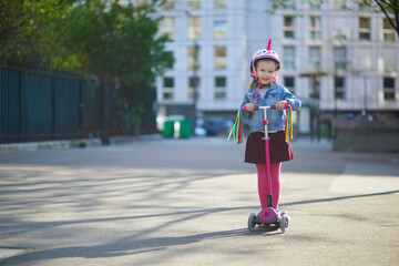 Adorable preschooler girl riding her scooter in a city park on sunny spring day