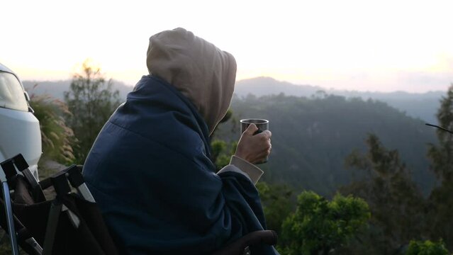 Lonely life of woman sitting with mug of coffee near camping trailer overlooking forest at dawn. Free life on camping trailer travel by wildlife landscape. Concept travel on camping trailer to retreat