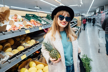 Stylish fashion smiling woman holding fresh organic pineapple in the supermarket store during selecting fresh products. Grocery Shopping. Healthy eating diet, go vegan. Selective focus. Copy space.