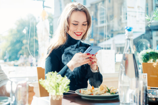 Cheerful young woman taking picture of her lunch while sitting at the cafe table. Woman use phone to take photo of her meal. Food Blogger, influencer making video for social networks. Selective focus.