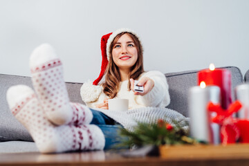 Relaxed, smiling woman in santa hat, jumper and warm socks sitting on comfortable couch, using tv...