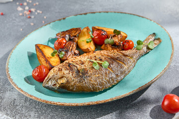 A side perspective of a plated grilled Dorada fish, garnished with potatoes, mushrooms, and cherry tomatoes. Served on a blue ceramic plate for a wholesome meal