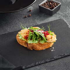 Elegant bruschetta topped with shrimp, fresh greens, avocado spread, and vibrant red garnish, set on a dark slate beside peppercorns