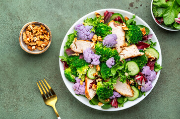 Healthy fresh salad from purple cauliflower, broccoli, grilled chicken breast, cucumbers, red onion, radicchio, spinach with walnuts, green table background, top view