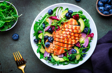 Fresh salad with blueberries, grilled chicken breast, avocado, red onion, walnuts and arugula on green table background, top view