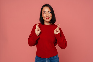 Beautiful asian woman holding fingers crossed while standing isolated over pink wall