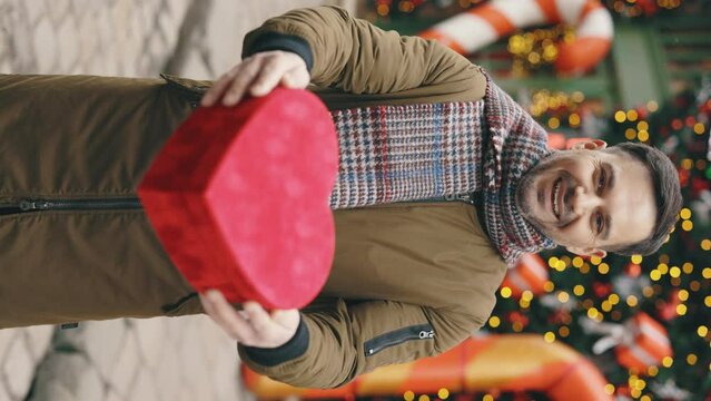 Vertical video. Caucasian bearded man standing in the winter decorated street holding the valentino heart shape gift. Handsome man outside with the red christmas present.