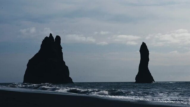 Huge sea stacks of the famous Black Beach in Iceland. Reynisfjara Black Sand Beach. Waves crash on the rocks and seagulls fly around the majestic rocks. Still shot in slow motion, 4k.
