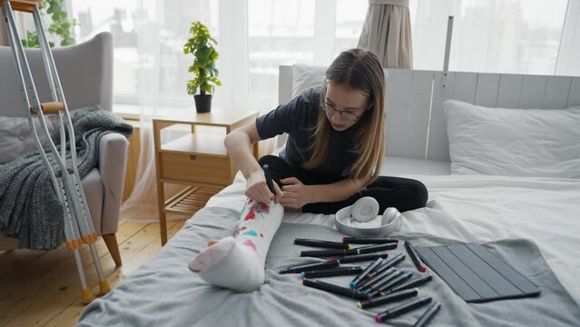 Teen girl painting colorful pictures using felt-tip pens on orthopedic cast on broken leg sitting in bed at home. Boring teenager resting relaxing drawing. Recovery of damaged leg, sick leave concept.