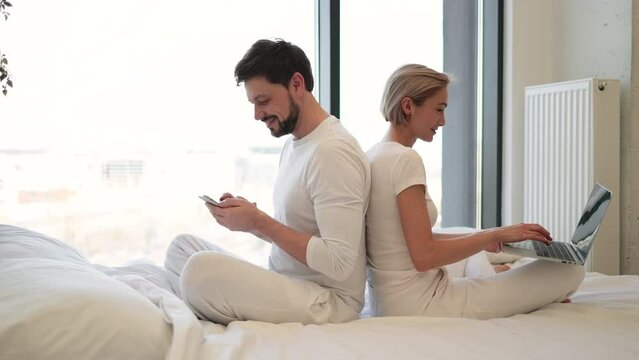 Young woman sitting in bed and sending text messages using modern laptop while her husband sitting back to her back with smartphone. Loving married couple achieving tech-life balance resting at home.