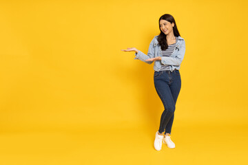 Happy Asian woman traveler presenting or showing open hand palm with copy space for product and wearing jeans jacket isolated over yellow background