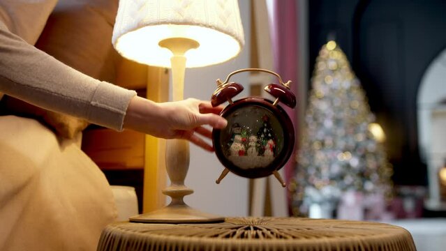 It's Christmas time! Alarm clock with snow inside near the bed on Christmas night against the backdrop of a beautiful decorated Christmas tree in the living room in the evening. Christmas night and ma - Powered by Adobe
