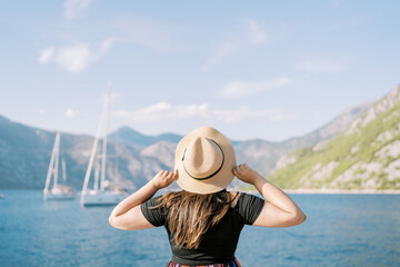 Woman stands on the shore holding a straw hat on her head and looks at the sailboats. Back view
