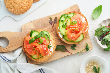 Open smoked salmon sandwiches with cream cheese, cucumber, sesame seeds, microgreens, spinach, and peas leaves on light old wooden background. Healthy breakfast food. Delicious snack. Top view.