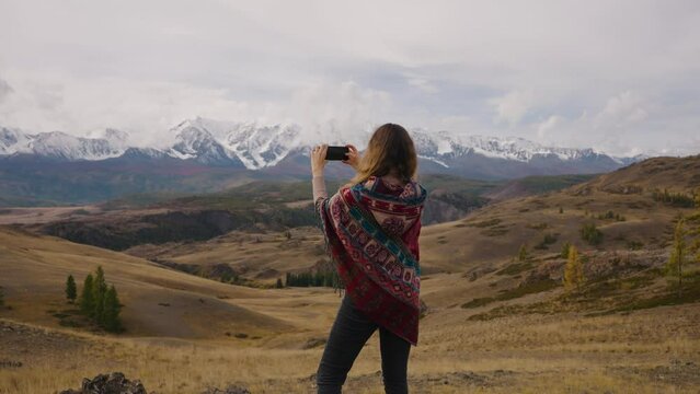 A female traveler takes pictures on her phone of a stunning landscape of snowy mountains during her car trip