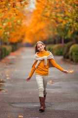 a teenage girl in a yellow sweater walks along a street outside the city against the backdrop of yellow trees. fun walk in autumn
